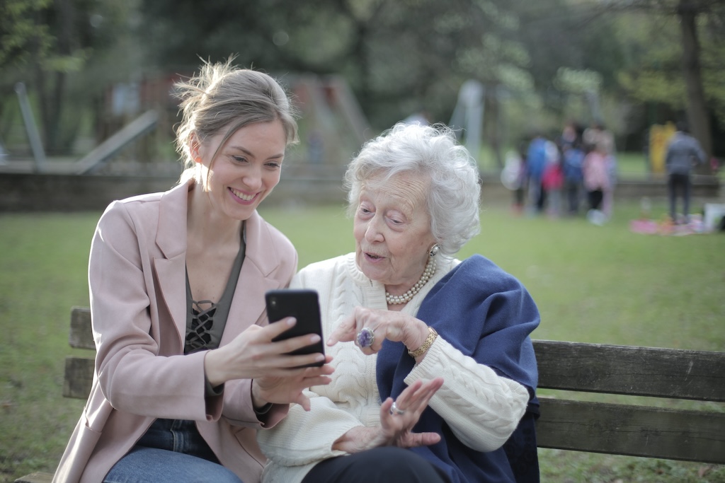 Two people sitting on a bench in a park, looking at a phone together.